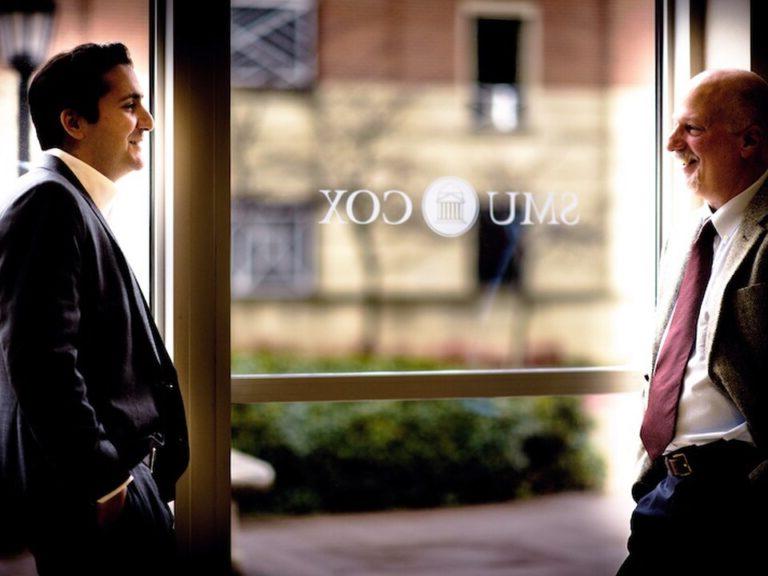 Two SMU Cox faculty members have a conversation in front of a window that has the school emblem printed in white